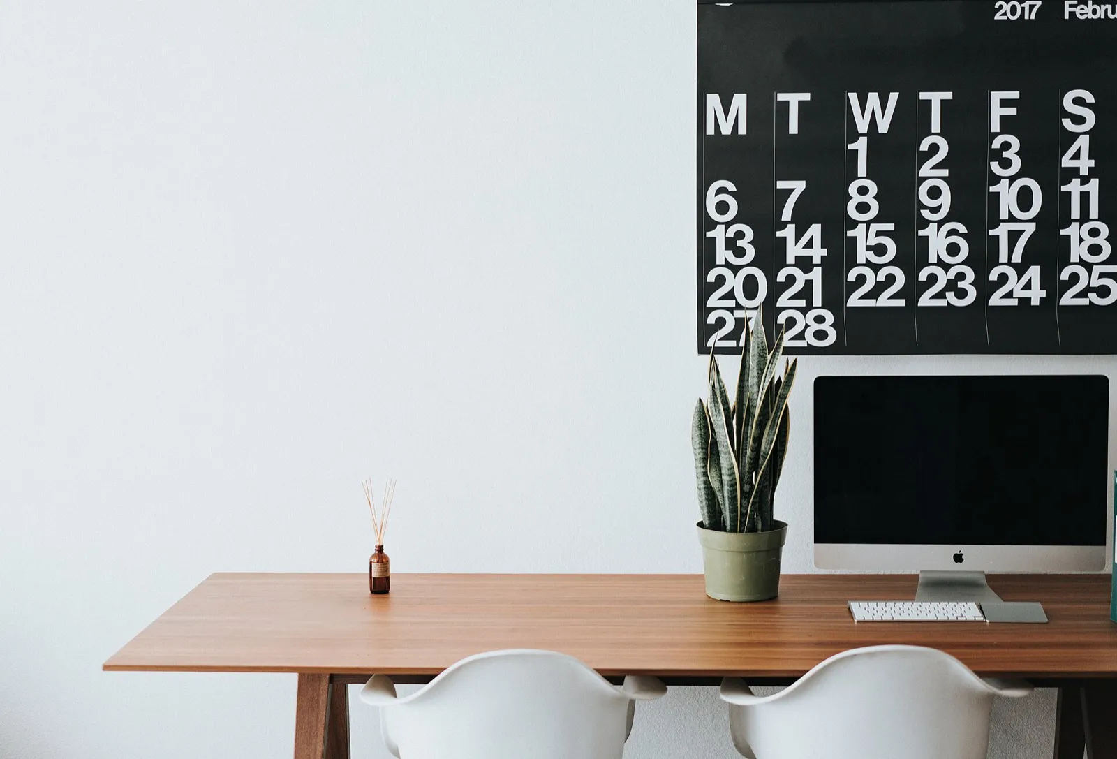 A clear wooden desk with a computer and a large month calendar on the wall